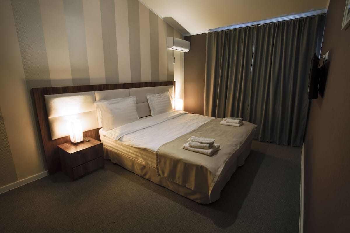 A hotel room with a neatly made bed, a bedside lamp, a wall-mounted air conditioner, and a window with curtains.