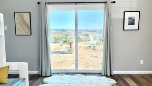 Una habitación con un amplio ventanal corredero que da a un paisaje seco. En el suelo hay una alfombra blanca y mullida, una silla blanca con un cojín amarillo, y dos cuadros en las paredes.