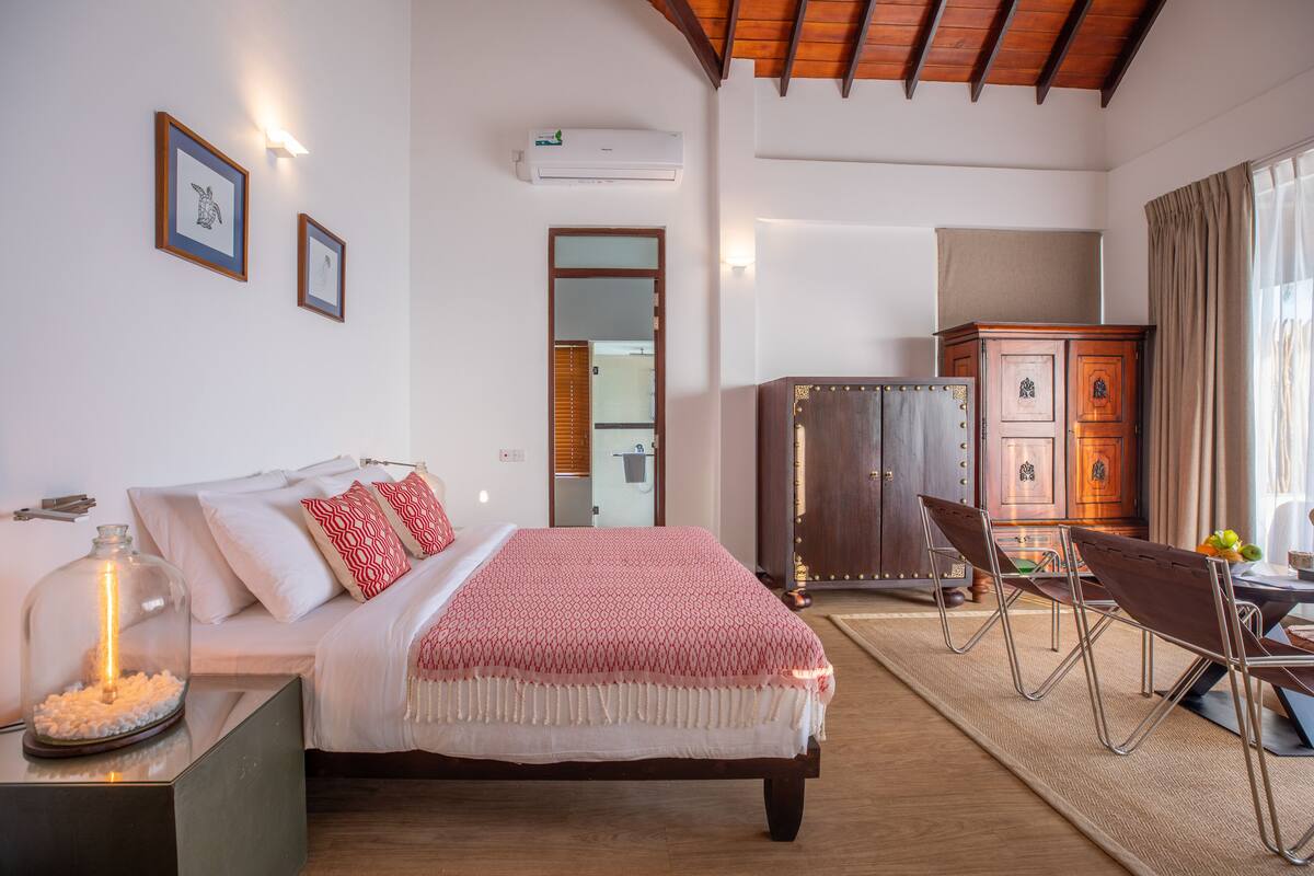 A bedroom with a bed, bedside table, and a mirror. There is a wooden cabinet and a dining area with chairs.