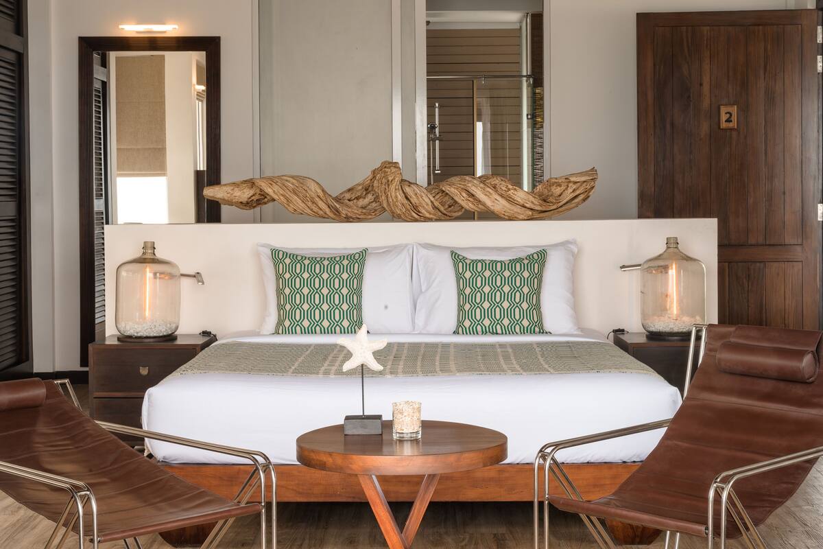 A modern hotel room with a large bed, two bedside tables with lamps, a round coffee table, and two leather chairs.