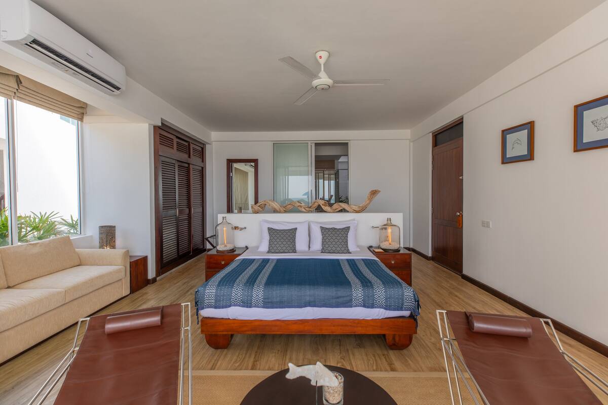 A bedroom with a large bed, a ceiling fan, a nightstand with a lamp, and a window with a view of the outdoors.
