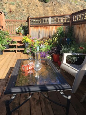 Une table à plateau en verre, entourée de chaises, est posée sur une terrasse en bois, elle-même bordée de plantes en pot et d’une clôture en bois.