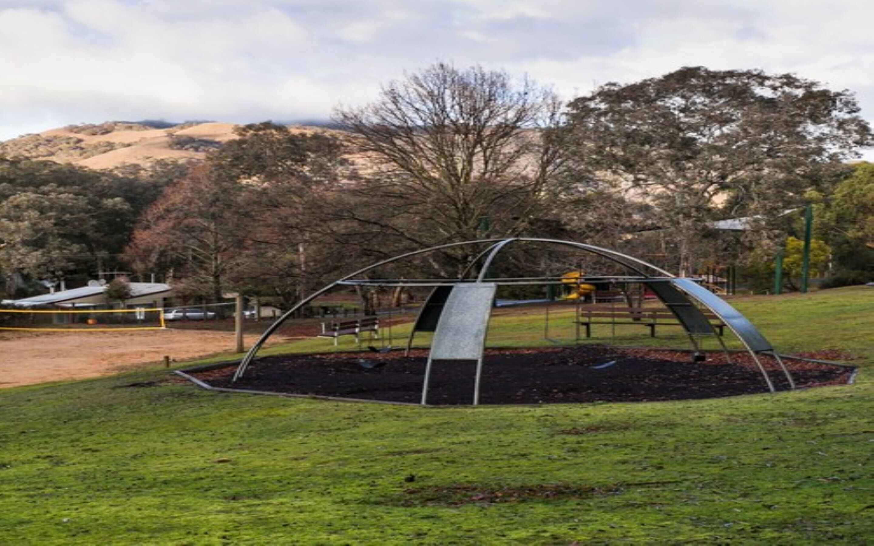 A park with a metal arch structure, a playground, and a grassy area with trees in the background.