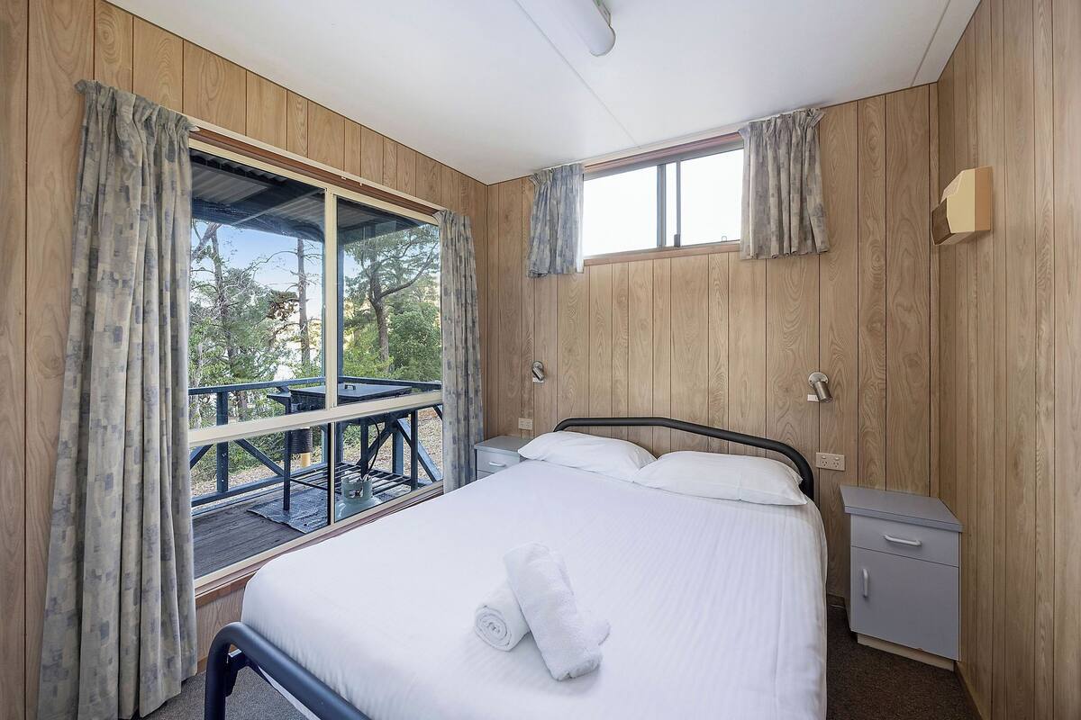A bedroom with a bed, a nightstand, a window with curtains, and a view of the outdoors.