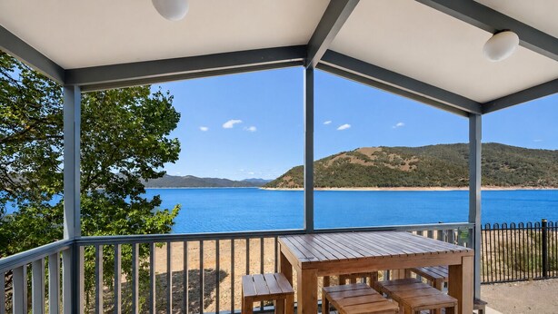 A covered outdoor area with a wooden table and benches, overlooking a clear blue sea and distant hills. - Reflections Burrinjuck Waters - Holiday Park (Burrinjuck)