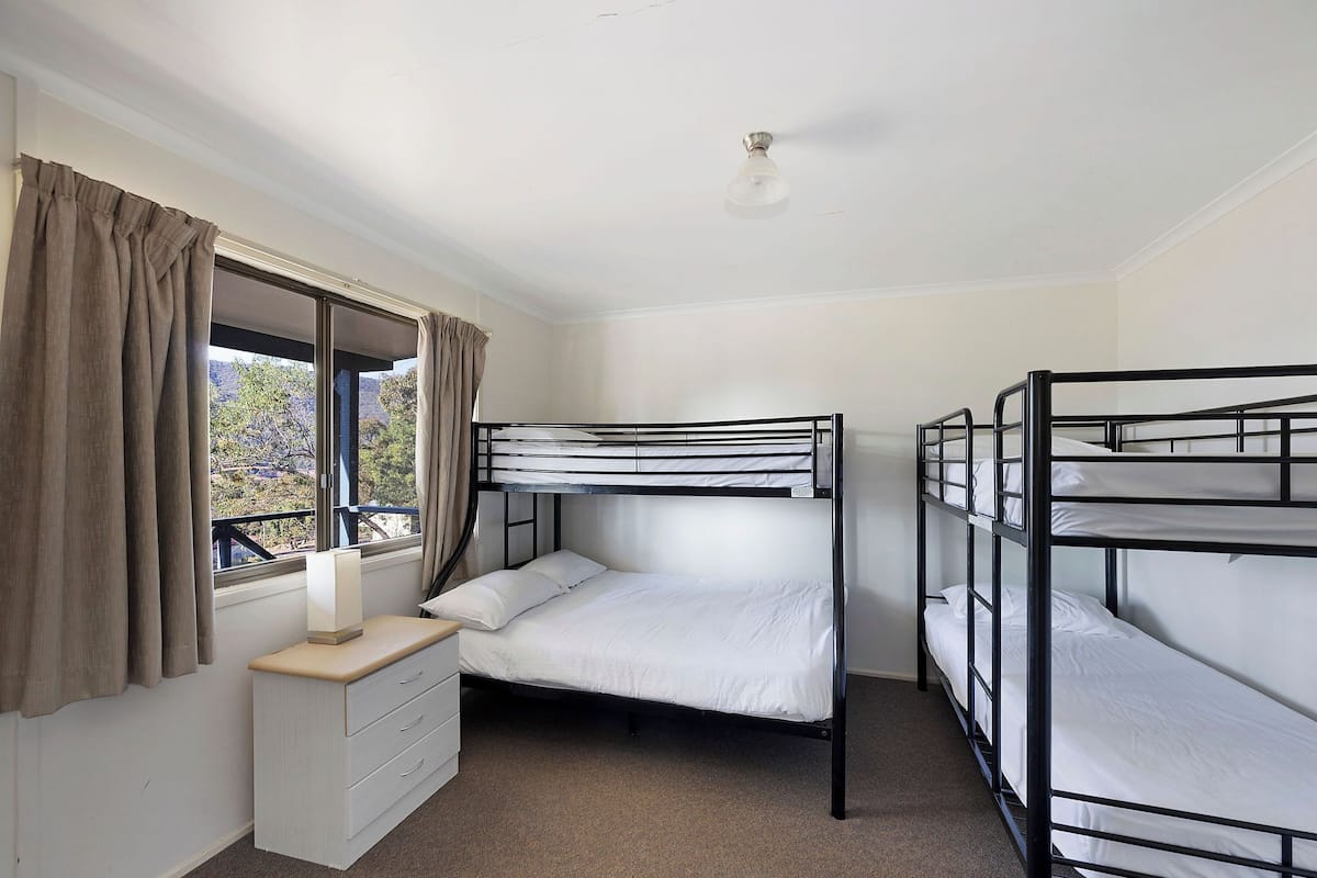 A room with bunk beds, a window with curtains, a bedside table with a lamp, and a ceiling fan.
