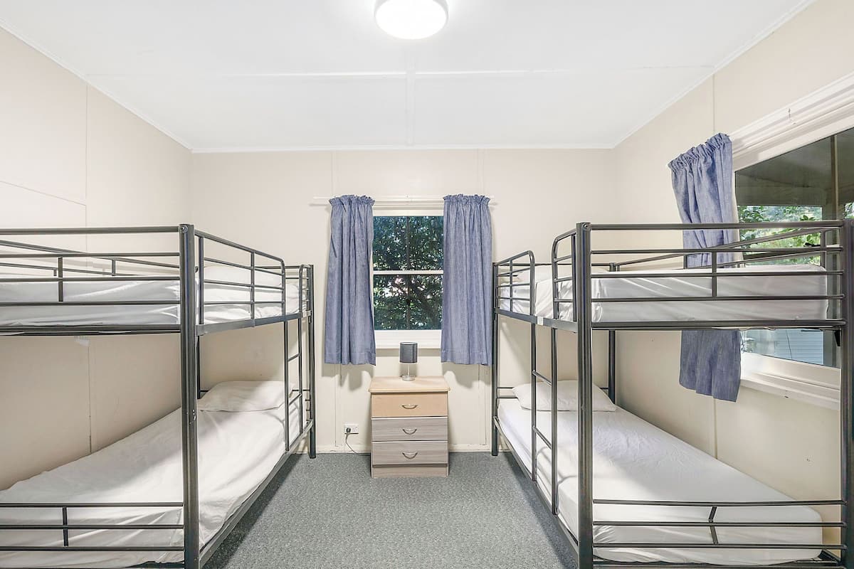 A dormitory room with bunk beds, a small bedside table, and a window with curtains.