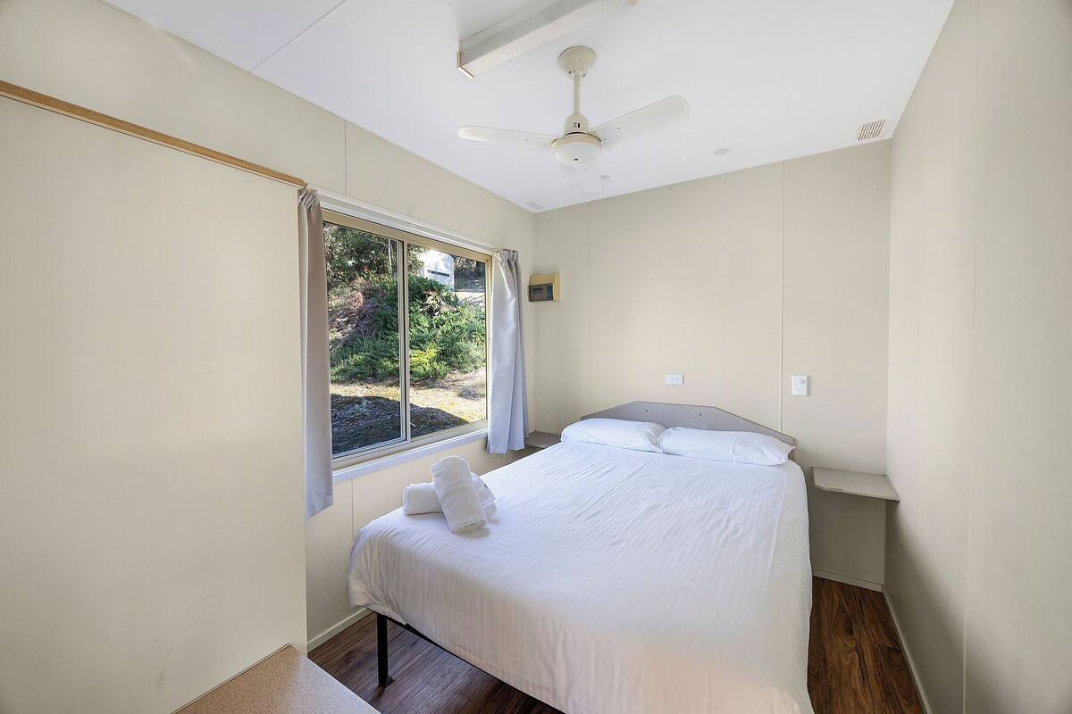 A bedroom with a bed, a ceiling fan, a window with curtains, and a view of greenery outside.
