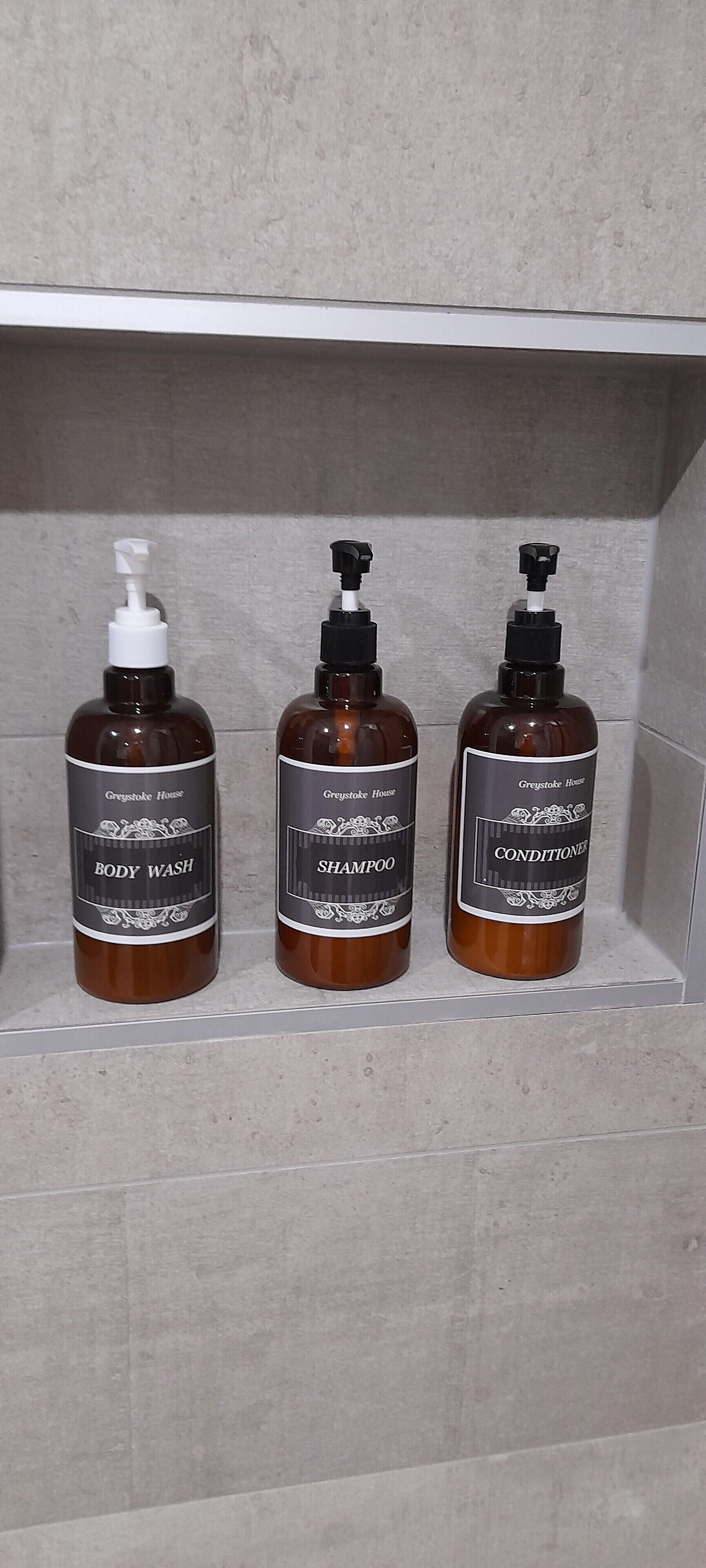 Tres botellas de jabón de cuerpo, champú y acondicionador Greystoke House sobre una estantería.