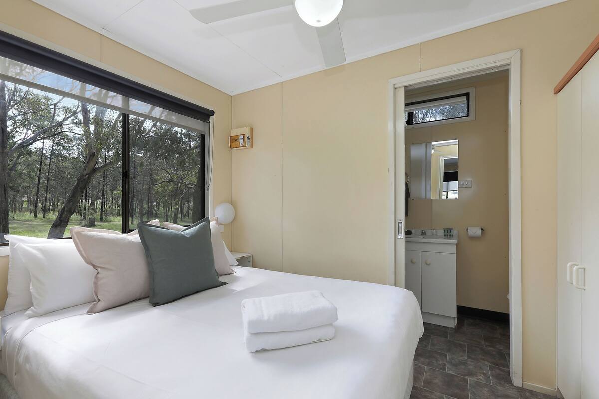 A bedroom with a large bed, a nightstand, a mirror, and a view of the outdoors.