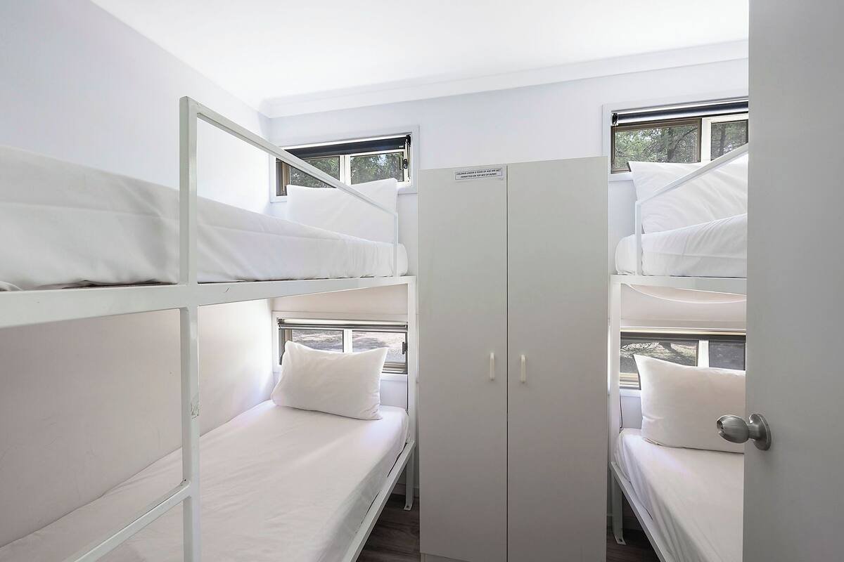 A dormitory room with bunk beds, a wardrobe, and windows with views of trees.