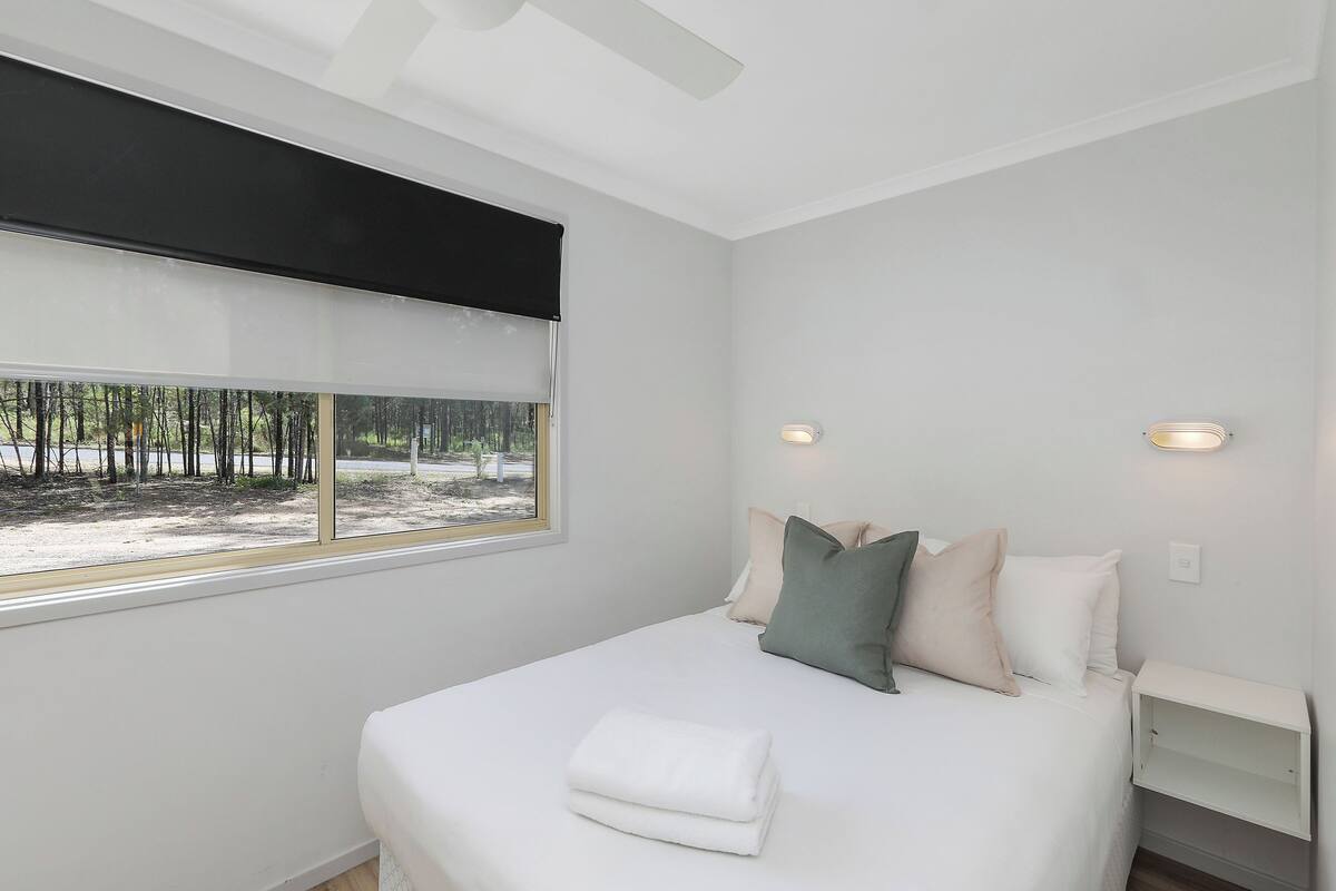 A bedroom with a bed, pillows, a bedside table, a window with a view of trees, and a ceiling fan.