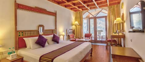 A hotel room with a wooden ceiling, a large bed with a headboard, a desk with a chair, and a television.