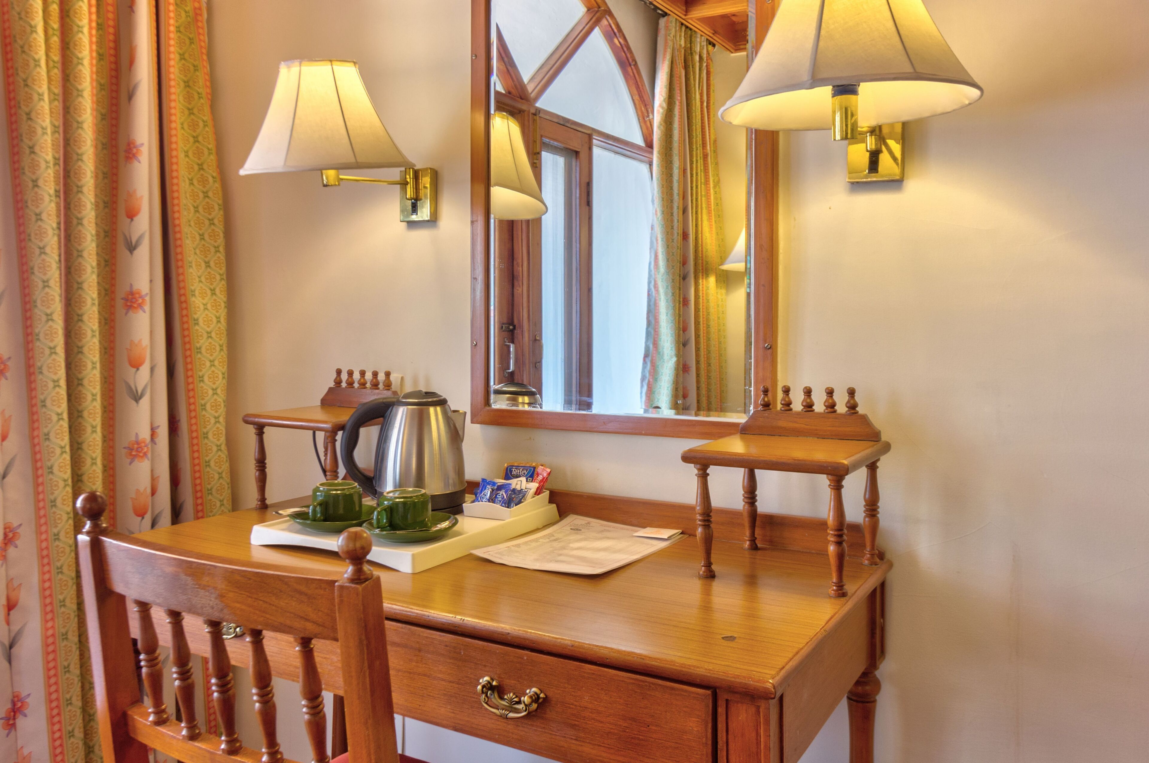 A wooden desk with a mirror, two wall-mounted lamps, a kettle, and a cup.
