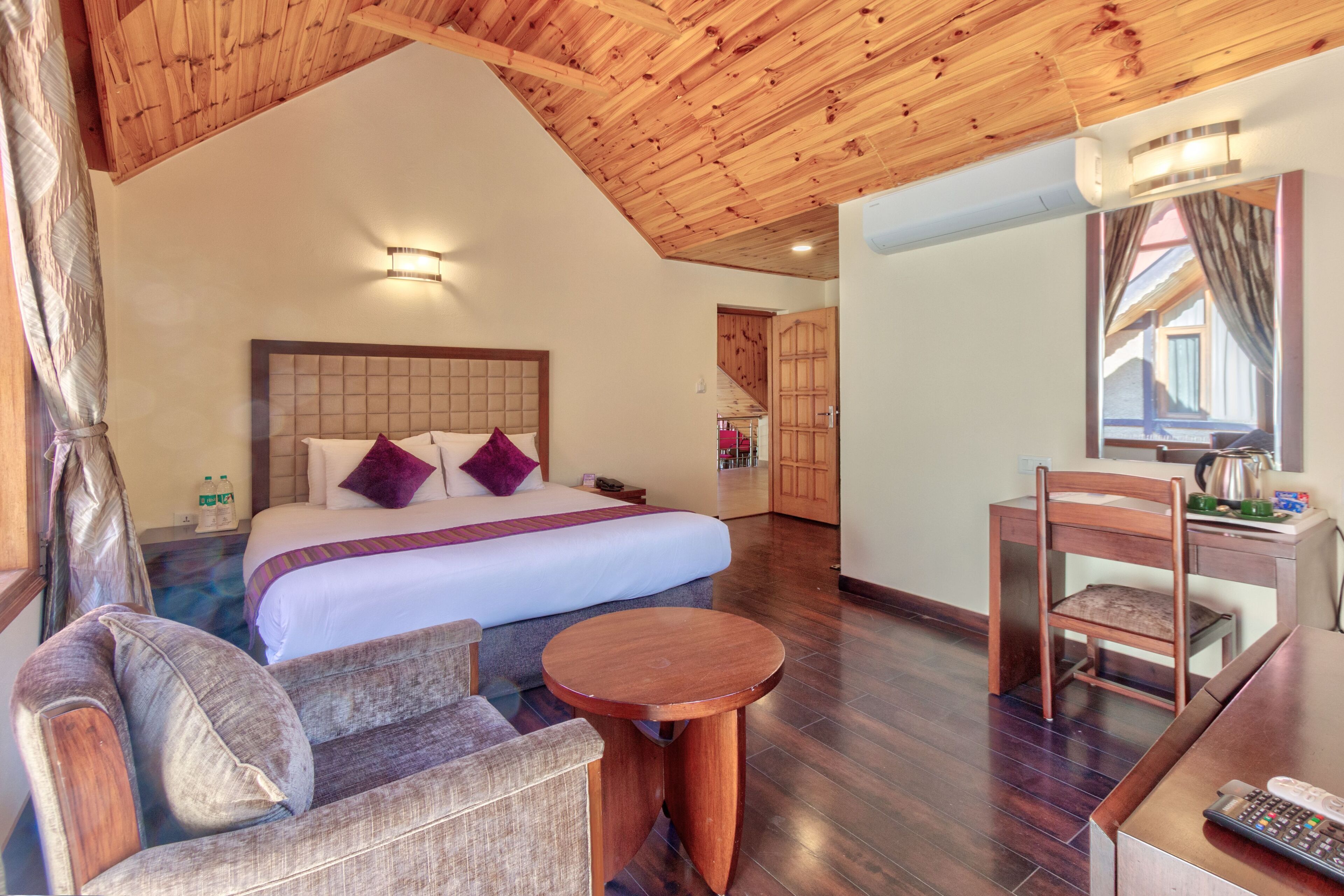 A bedroom with a large bed, a wooden ceiling, a desk with a chair, and a small round table.
