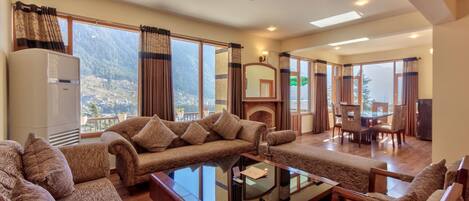 A spacious living room with a sofa set, a glass coffee table, and a fireplace. Large windows offer a view of the mountains.