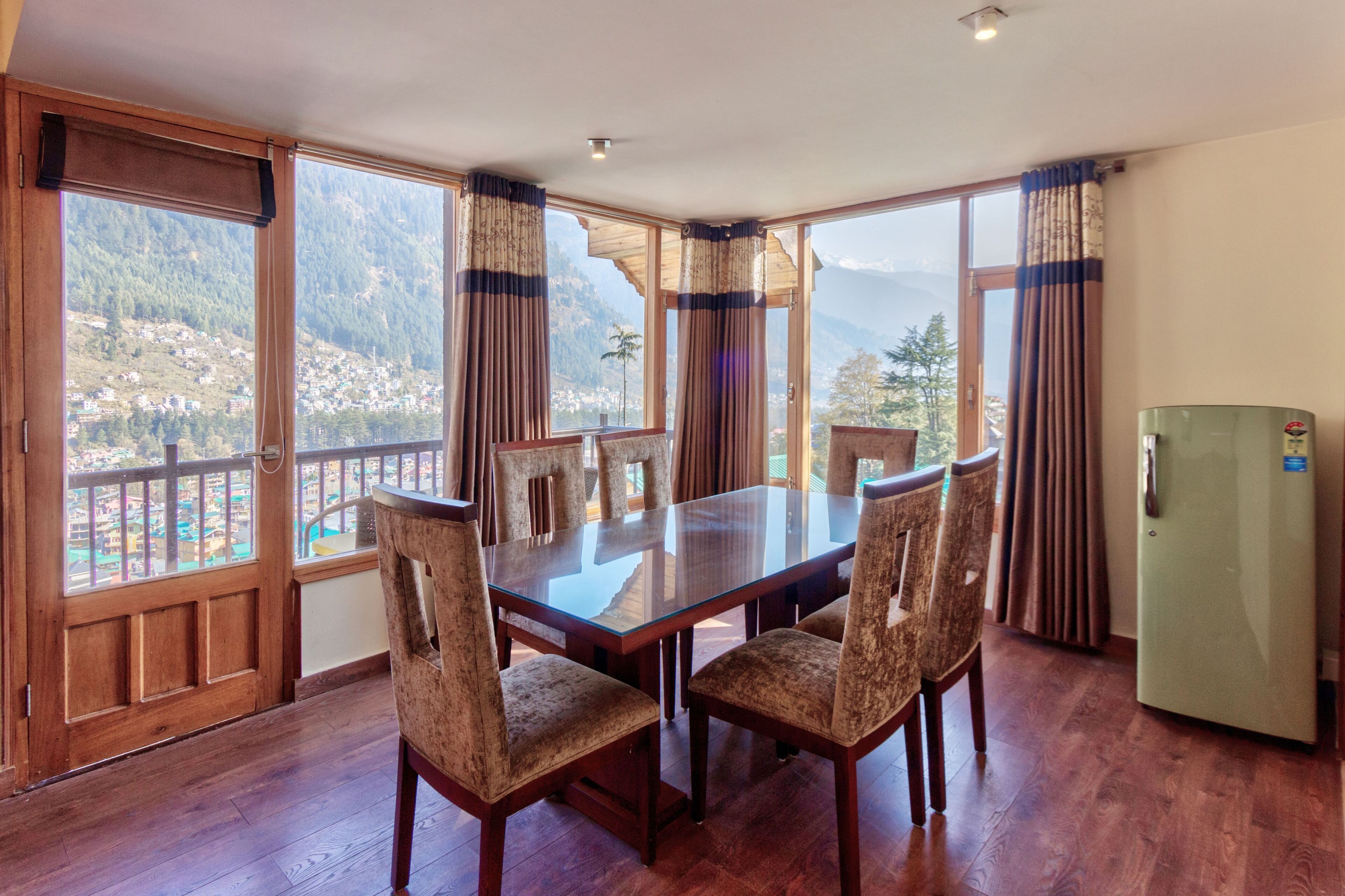 Un comedor con una mesa grande y cuatro sillas, ventanas amplias con cortinas y un refrigerador.
