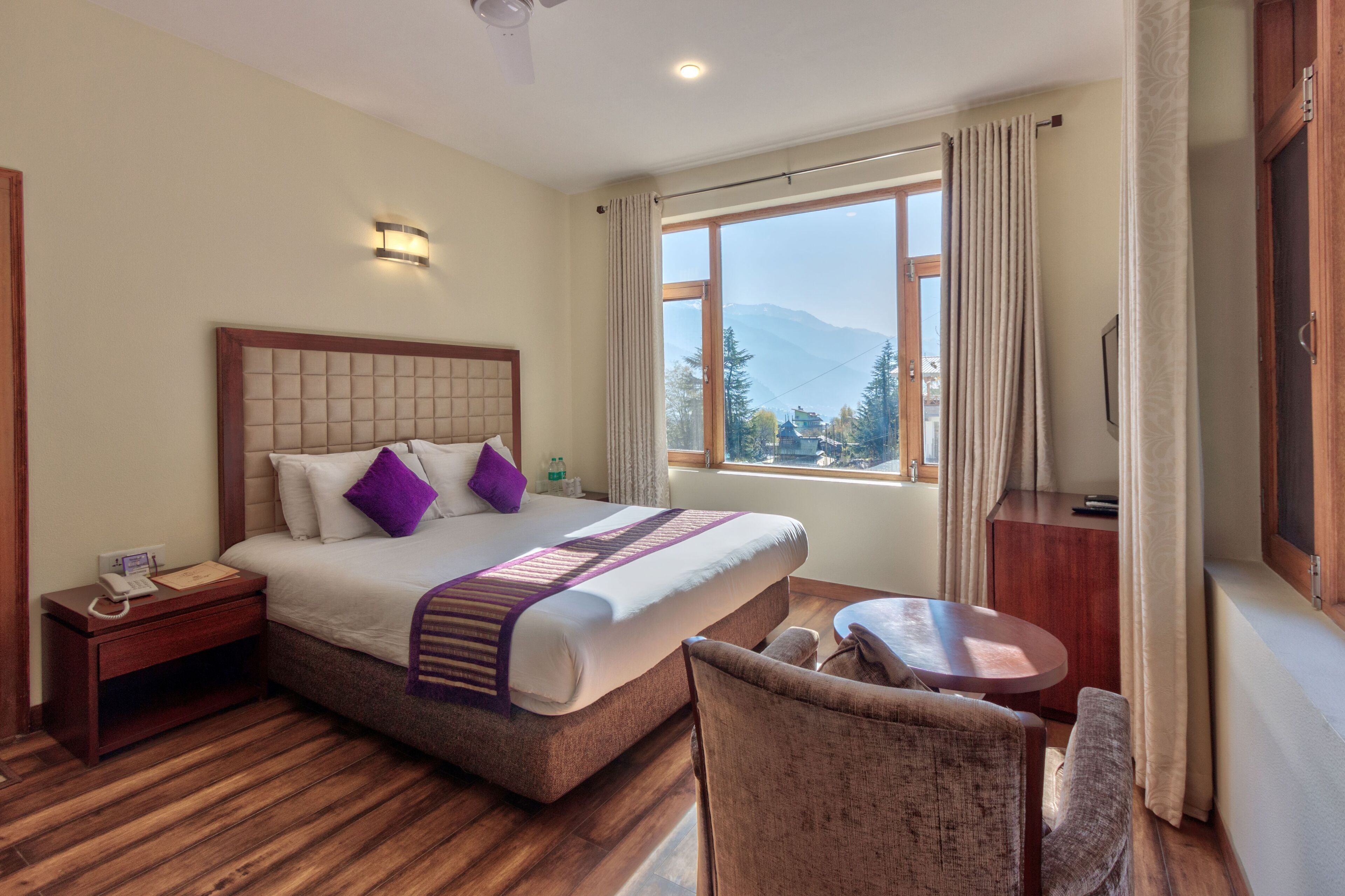 A hotel room with a large bed, a bedside table, a small table, a chair, and a window with a mountain view.