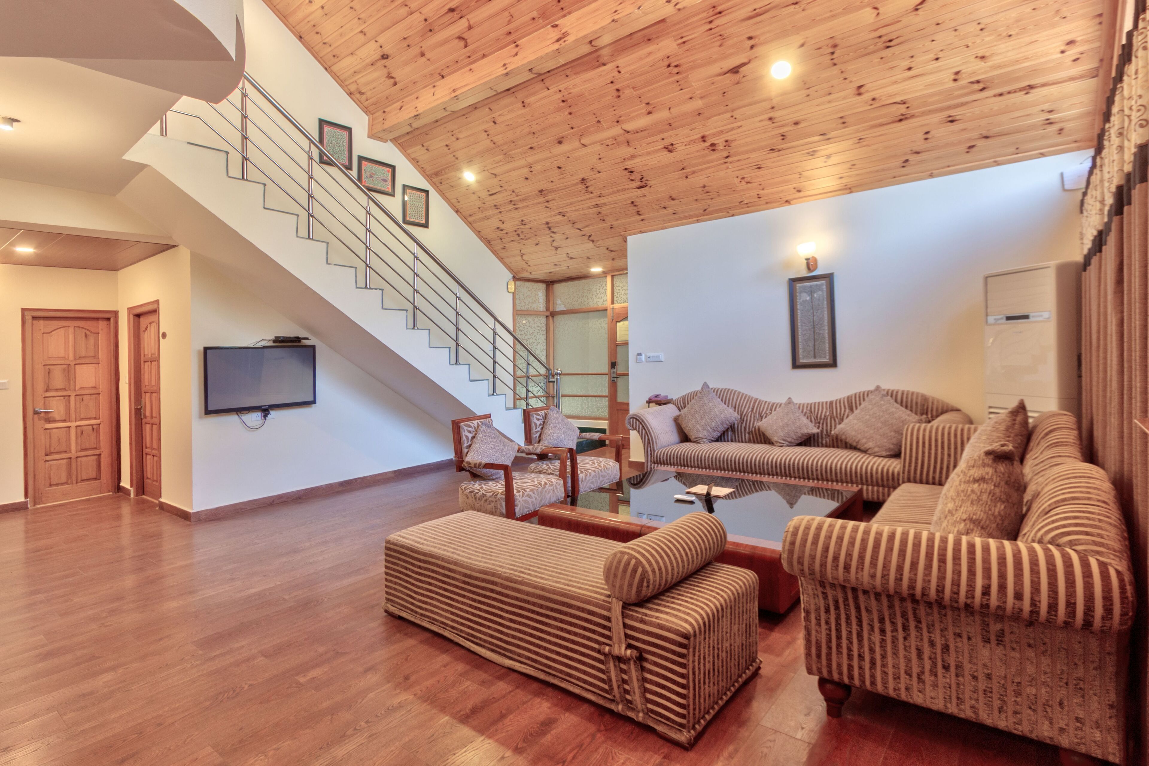 A spacious living area with a wooden staircase, multiple sofas, and a flat-screen TV mounted on the wall.