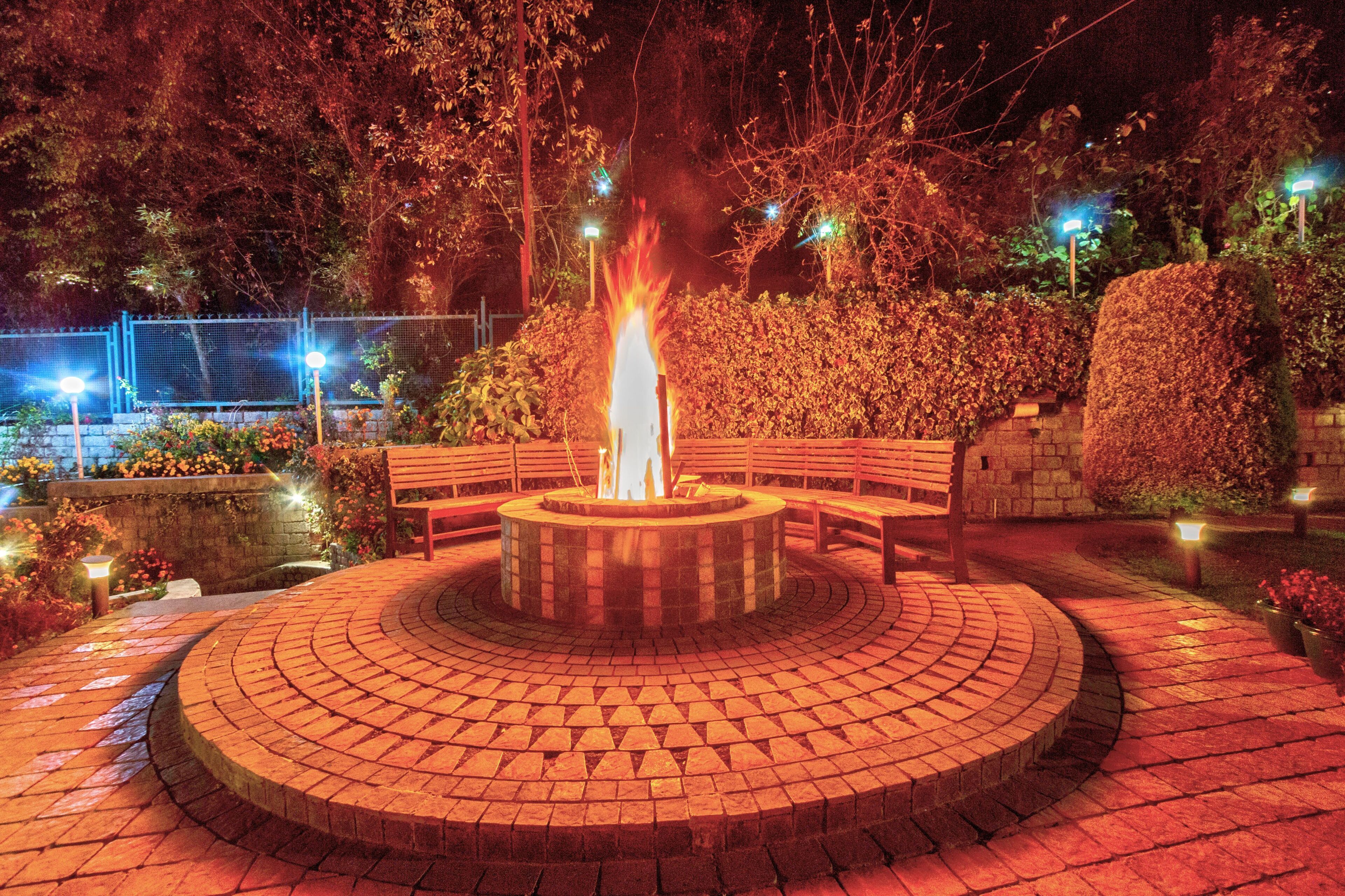 A circular brick fire pit with benches around it, surrounded by a brick pathway and greenery.