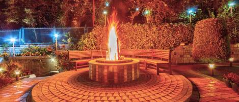 A circular brick fire pit with benches around it, surrounded by a brick pathway and greenery.