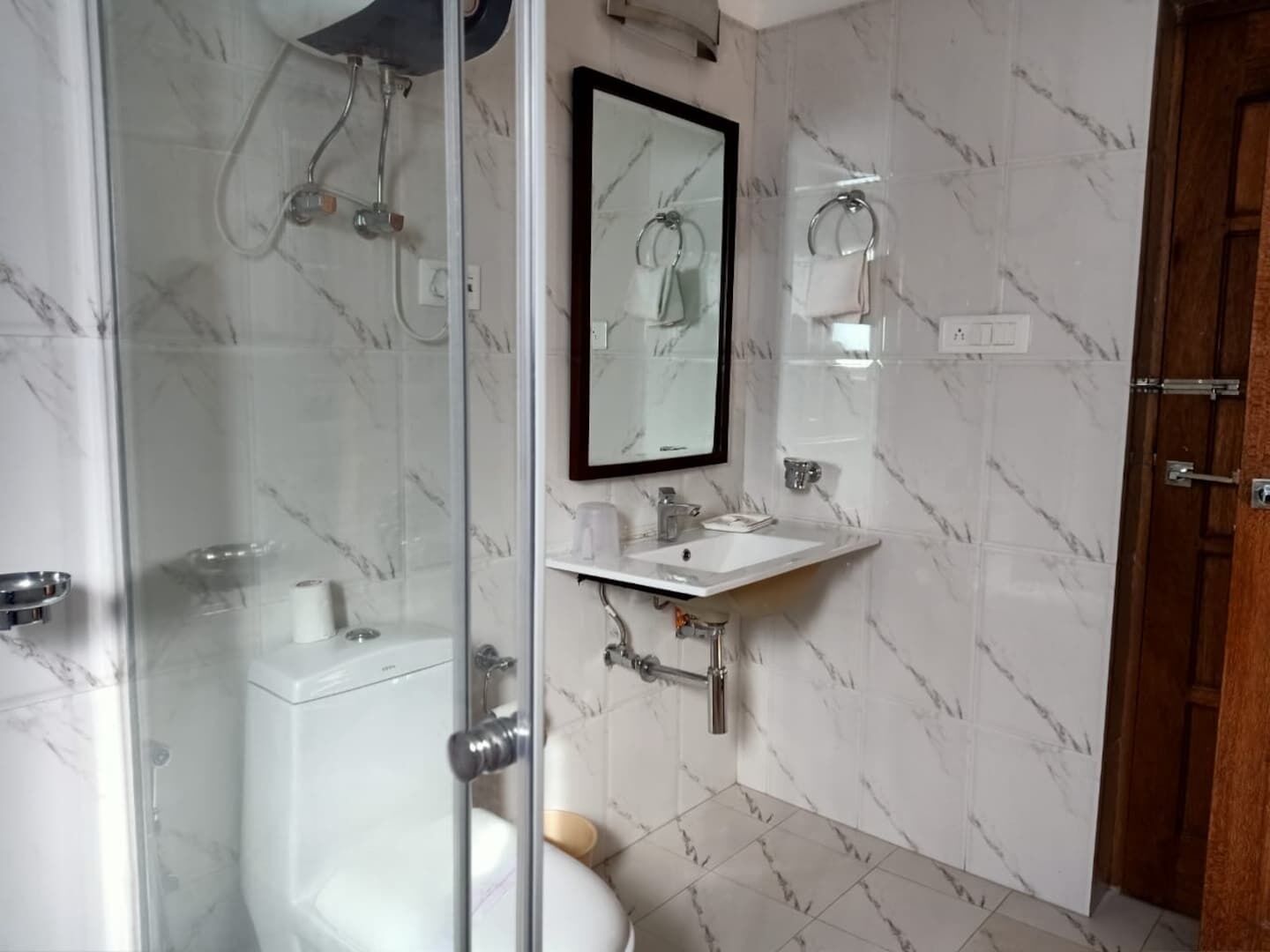 Une salle de bain dotée d’une douche à parois de verre, de toilettes, d’un lavabo avec un miroir au-dessus et de murs aux motifs de marbre.