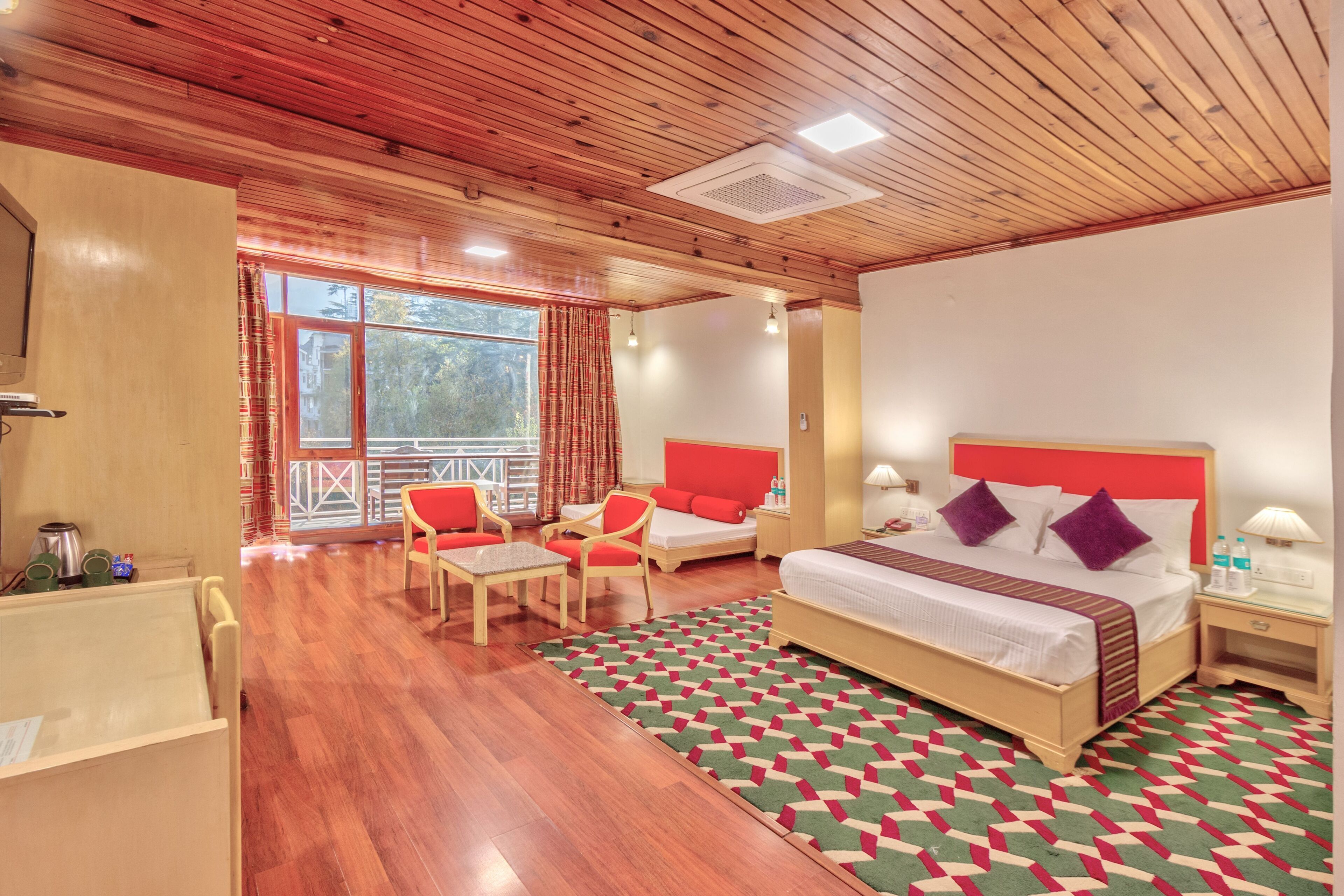 Ein Hotelzimmer mit Holzboden, einem großen Bett mit rot-weißer Bettwäsche, zwei Stühlen, einem kleinen Tisch und Blick ins Freie durch ein großes Fenster.