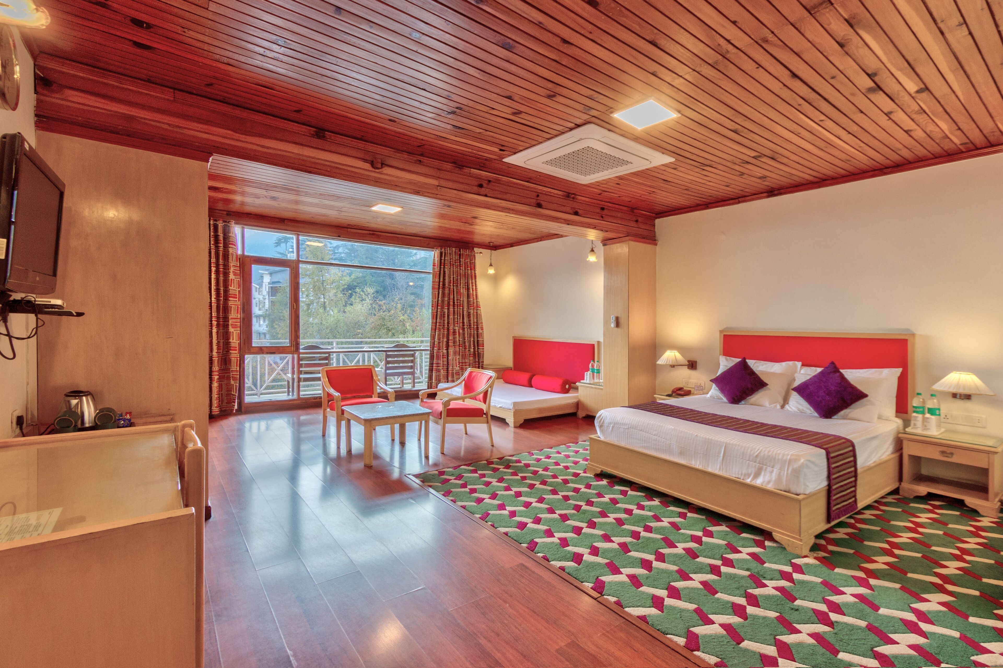 A hotel room with a wooden ceiling, a large bed with red and white bedding, a desk with a chair, a small table, and a view of the outdoors through a large window.