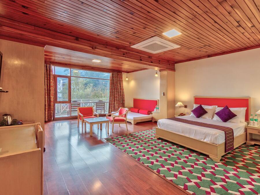 A hotel room with a wooden ceiling, a large bed with red and white bedding, a desk with a chair, a small table, and a view of the outdoors through a large window.