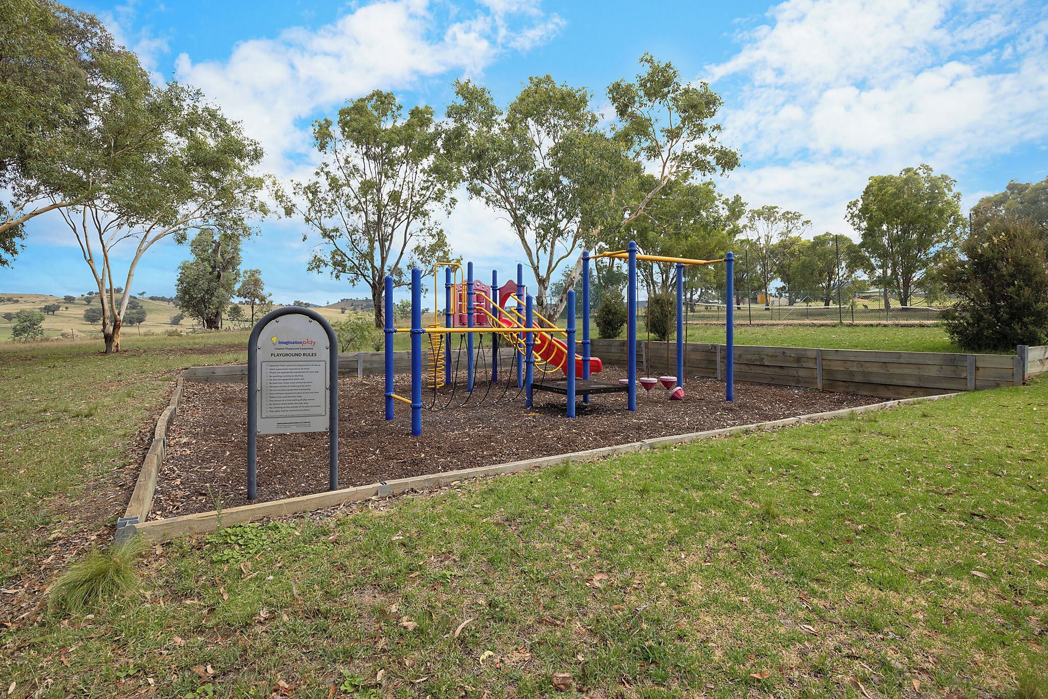 A playground with a slide and climbing structures, surrounded by trees and a grassy area.