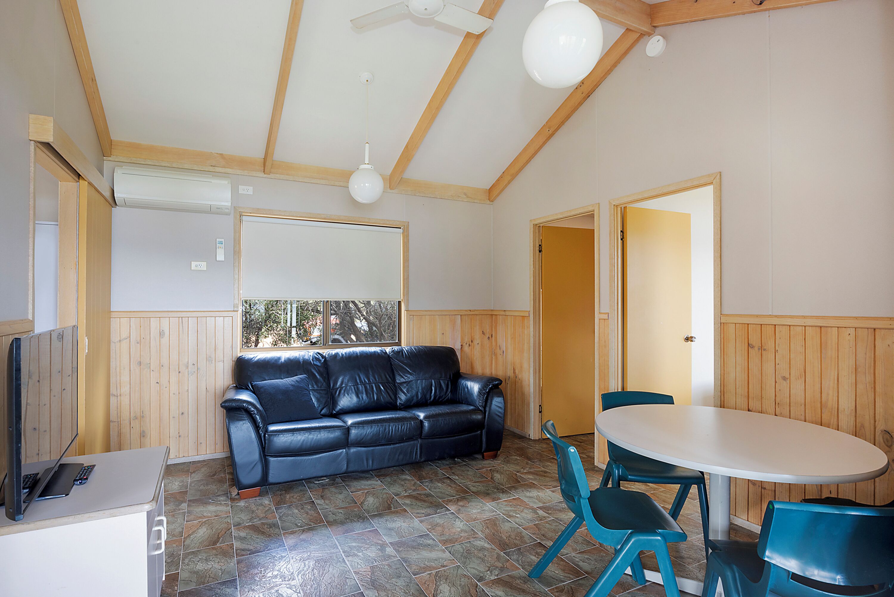 A room with a black leather sofa, a white table with blue chairs, a ceiling fan, and wooden walls.