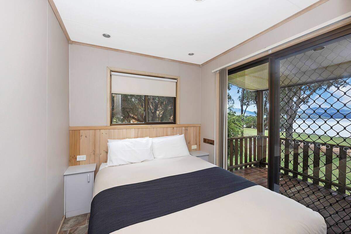A bedroom with a bed, a nightstand, a window with blinds, and a view of the outdoors through a glass door.