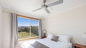 A bedroom with a large bed, a ceiling fan, a nightstand, a lamp, and a view of a grassy field through a window. - Reflections Grabine Lakeside - Holiday Park (Bigga)