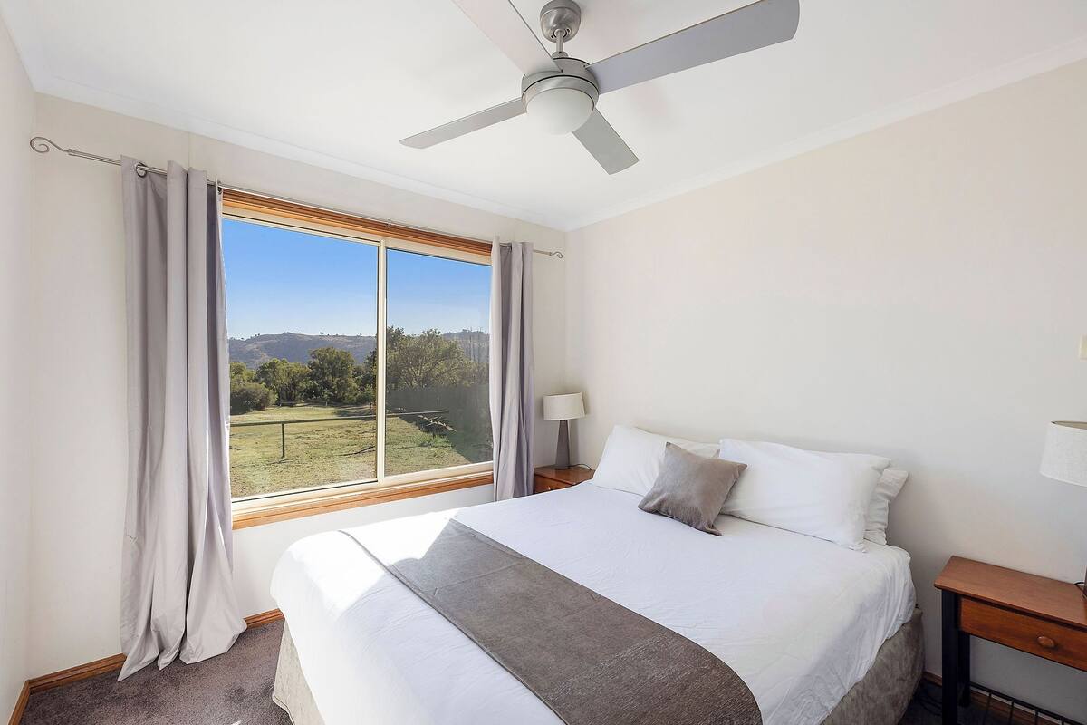 A bedroom with a large bed, a ceiling fan, a nightstand, a lamp, and a view of a grassy field through a window.