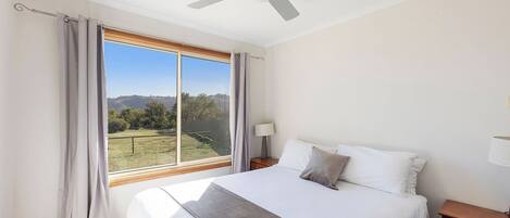 A bedroom with a large bed, a ceiling fan, a nightstand, a lamp, and a view of a grassy field through a window.