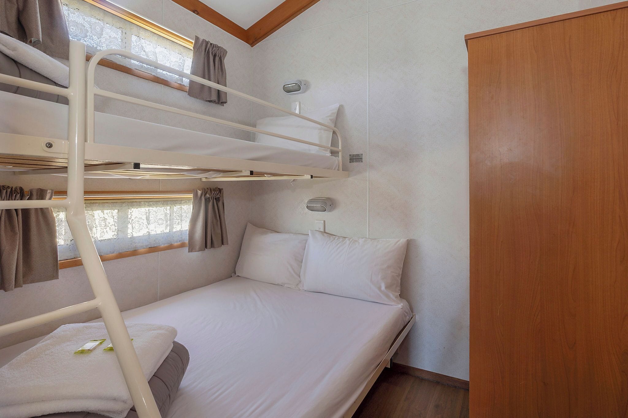 A bunk bed with white bedding and curtains, a wooden wardrobe, and a window with curtains.
