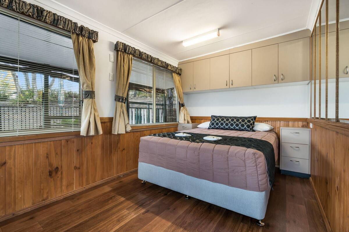 Ein Schlafzimmer mit Holzboden, einem Bett mit gemustertem Überwurf, einem Nachttisch, einem Kleiderschrank und einem Fenster mit Vorhängen.