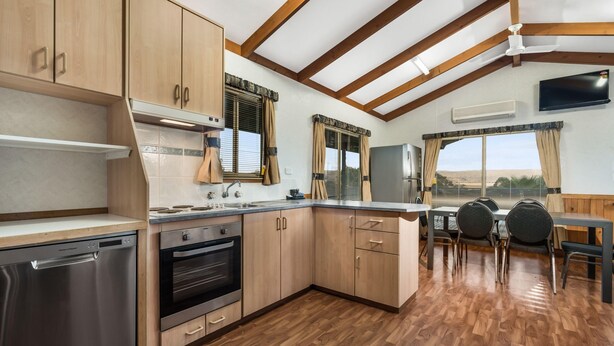 A kitchen with wooden cabinets, a dishwasher, an oven, and a dining area with a table and chairs. - Reflections Lake Burrendong - Holiday Park (Mumbil)