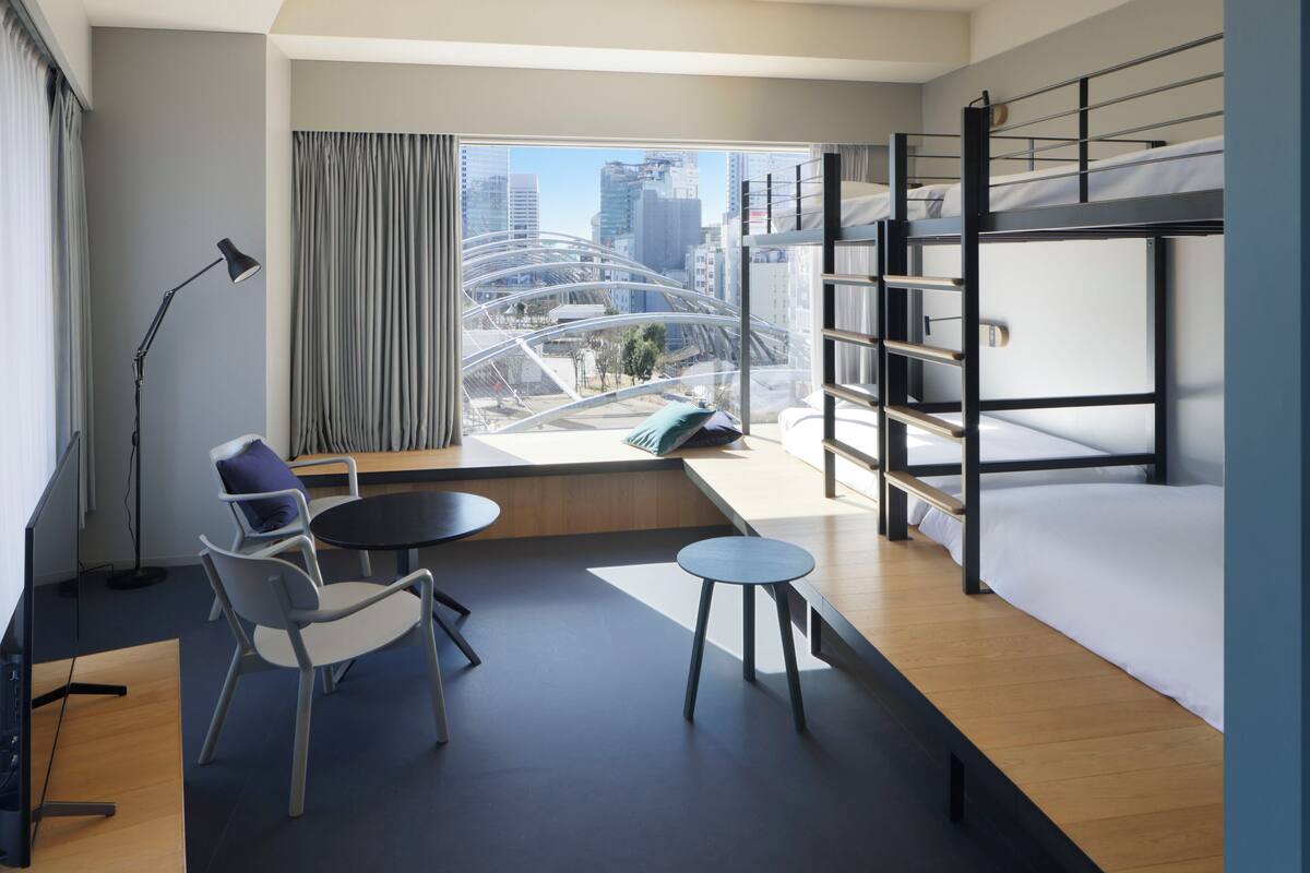 A modern hotel room with bunk beds, a desk, a chair, a small table, and a large window offering a city view.
