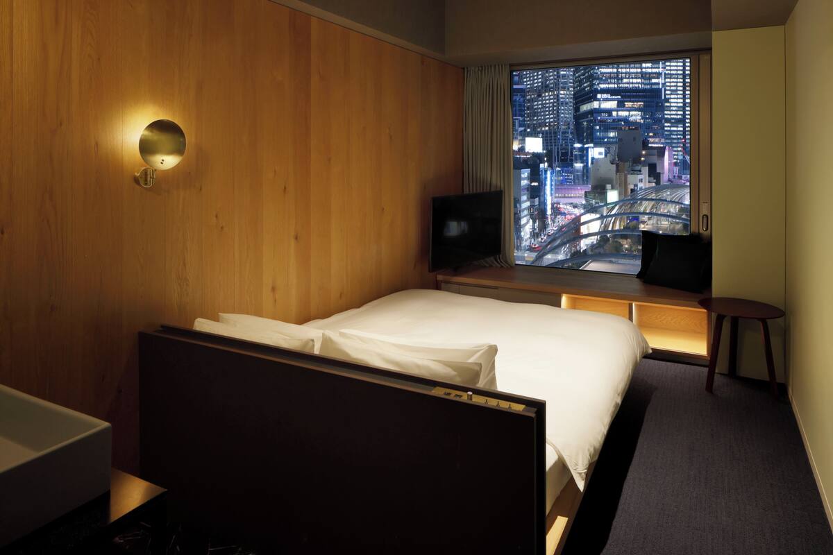 A hotel room with a bed, a TV, a small table, and a window with a city view.