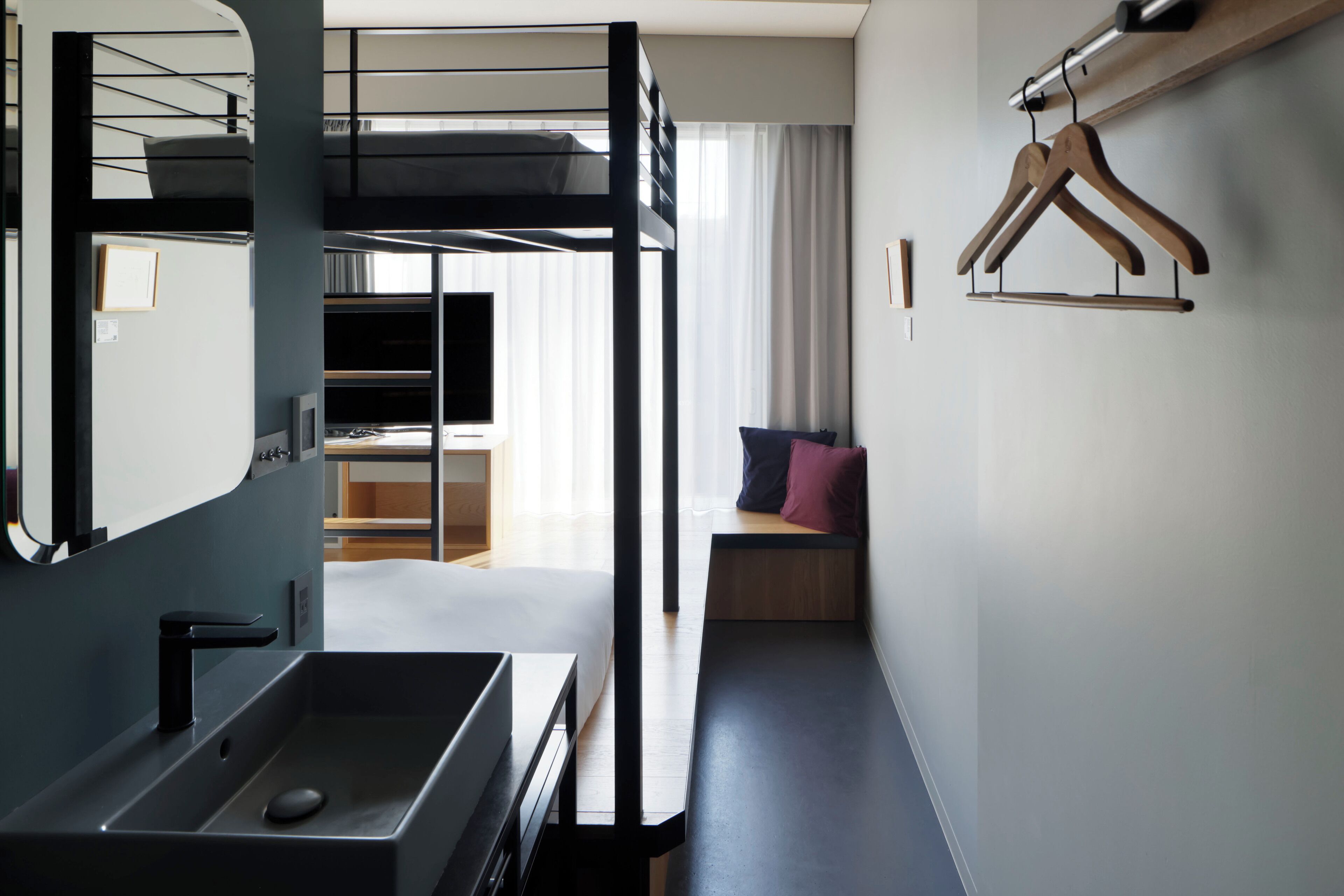 Habitación de hotel moderna con una cama individual superpuesta, un escritorio empotrado, un espejo grande y un banco de madera con cojines.