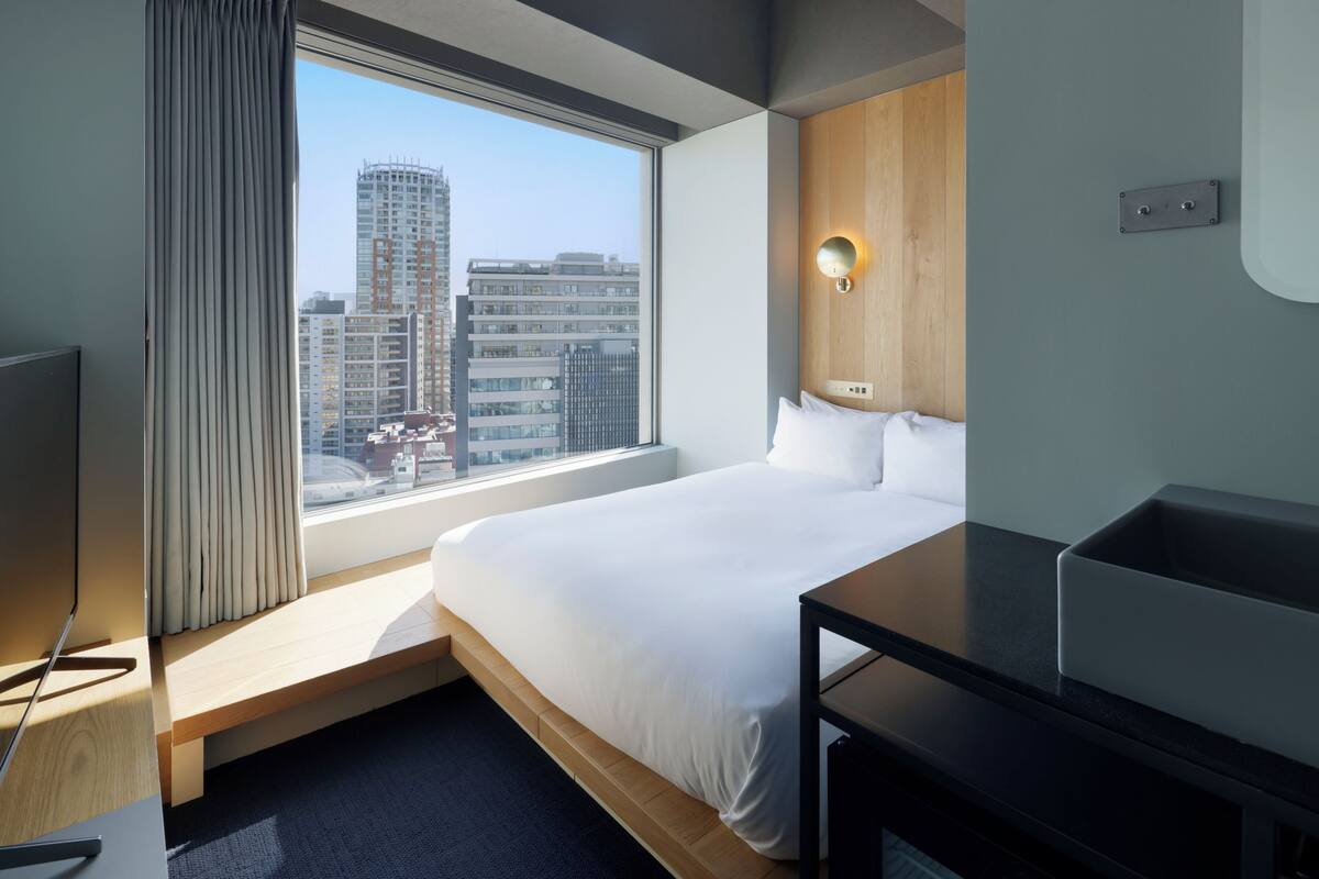 A hotel room with a large window, a bed, a desk, and a wall-mounted light fixture.