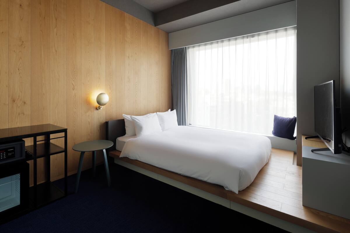 A hotel room with a wooden wall, a bed with white bedding, a small round table, a desk with a computer, and a television on a stand.