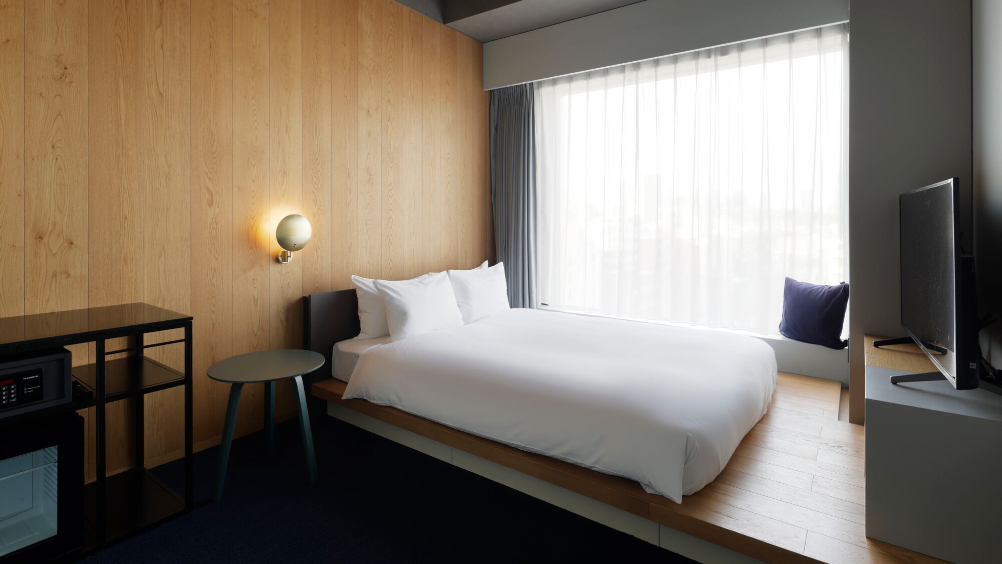 Una habitación de hotel con cama, escritorio, televisor y ventana con cortinas.