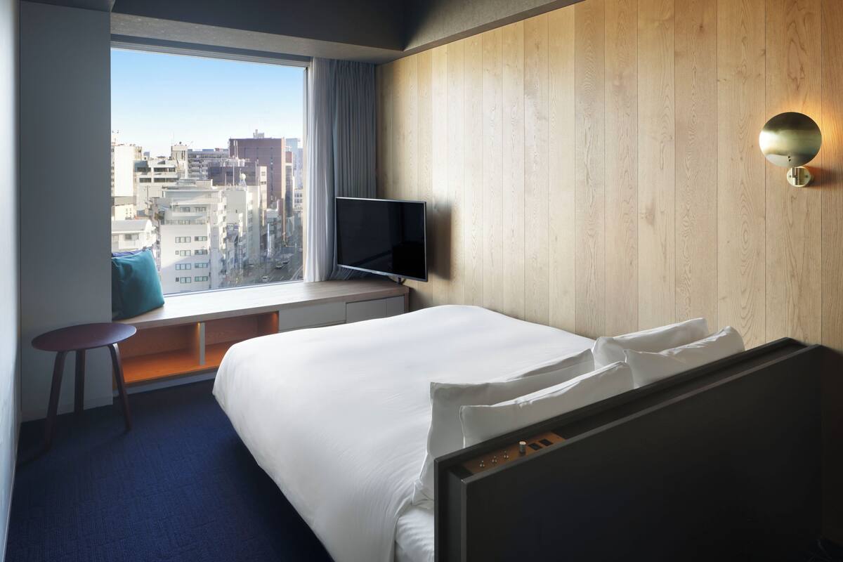 A hotel room with a large bed, a TV on a stand, a window with a view of buildings, and a wooden wall.