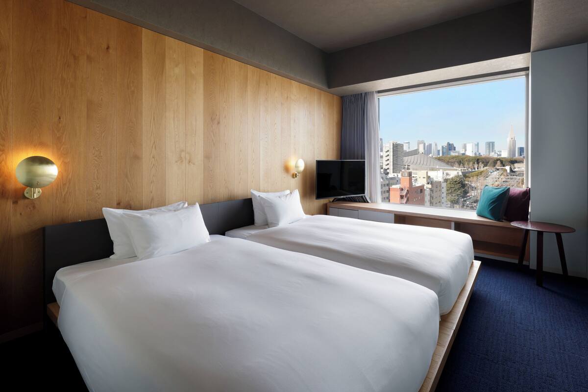 A hotel room with two beds, a TV, a window with a city view, and a small table with a lamp.