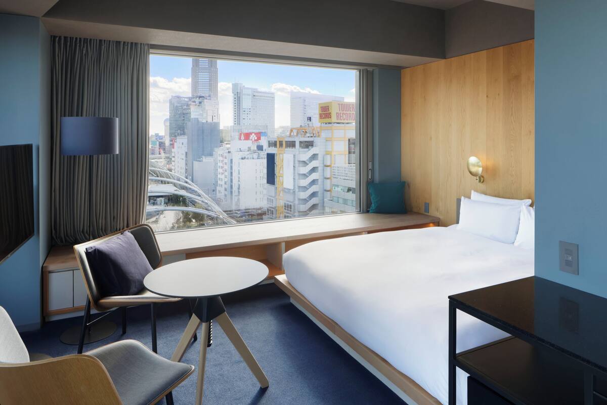 A hotel room with a large bed, a desk, a chair, a small table, and a view of the city through a large window.