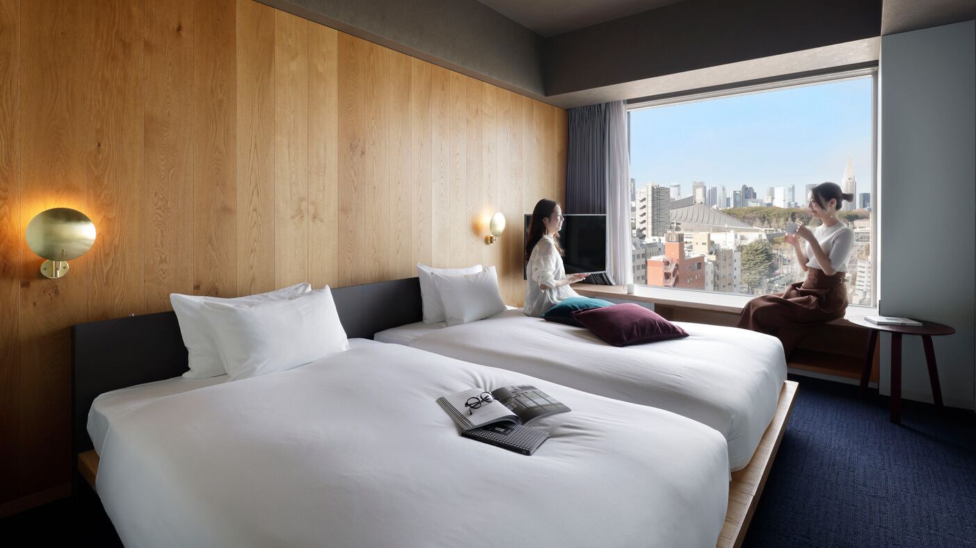 Habitación de hotel con dos camas, un ventanal con vista a la ciudad, una mesita pequeña y una lámpara fijada a la pared.