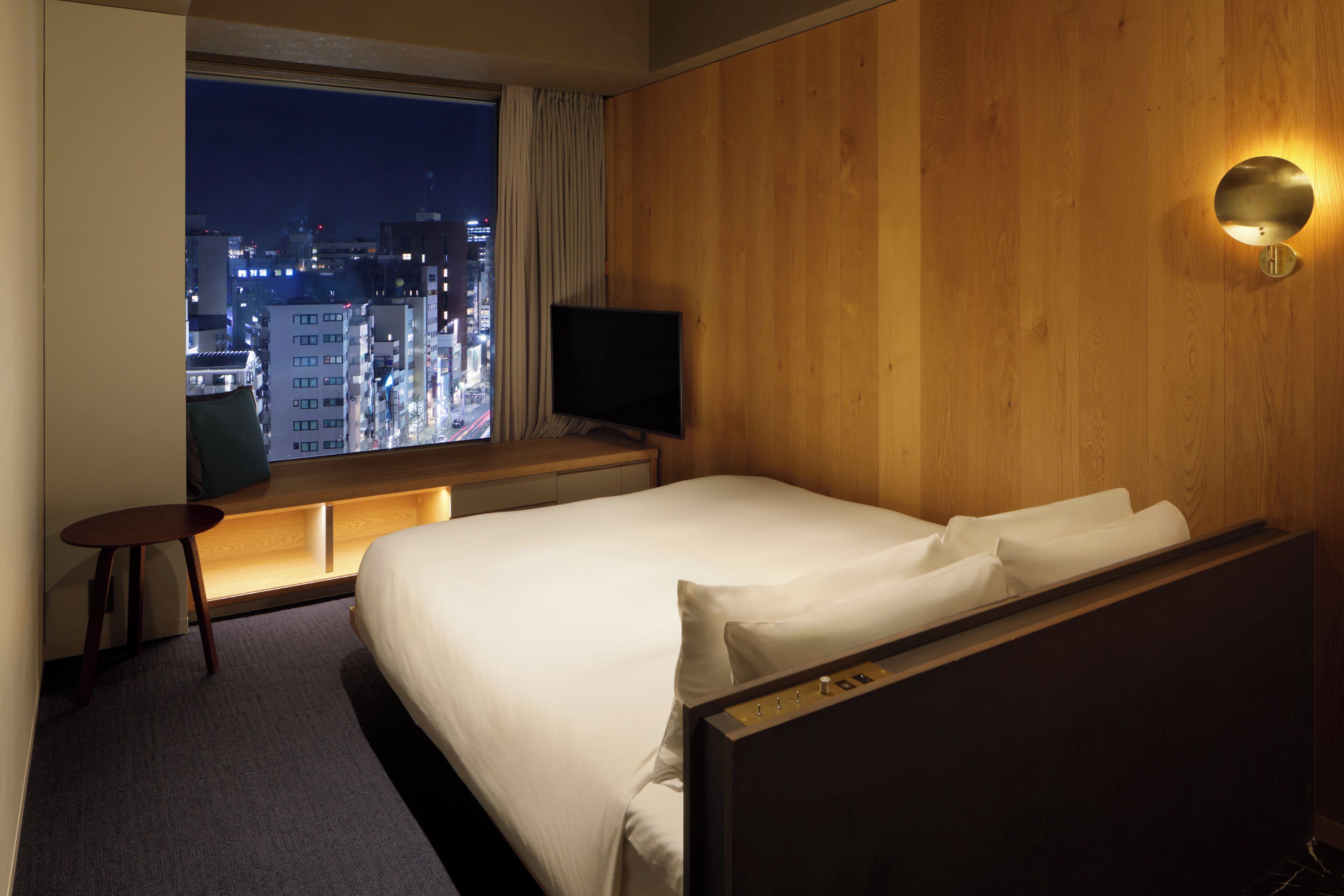 Habitación de hotel con una cama grande, televisión, pared de madera y ventana con vistas a la ciudad de noche.