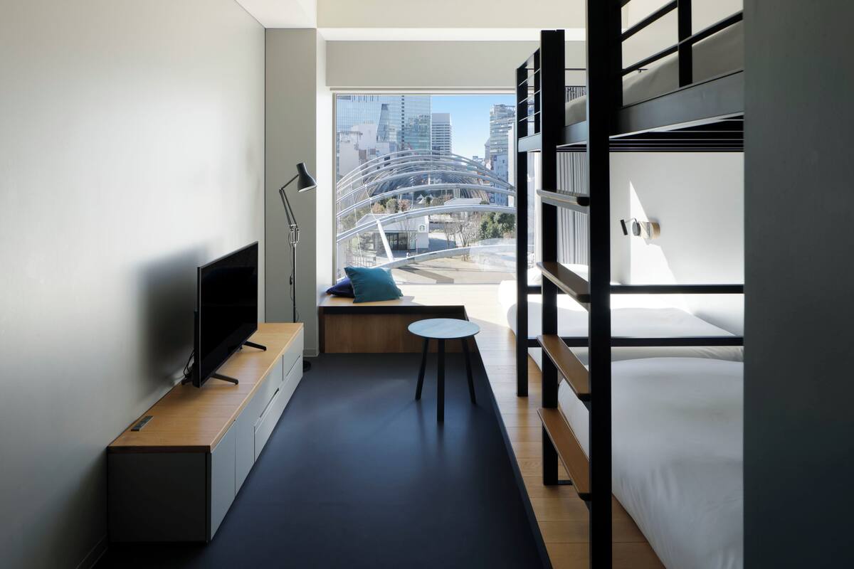 A modern hotel room with a TV, a small table, a bench with a pillow, and a bunk bed.